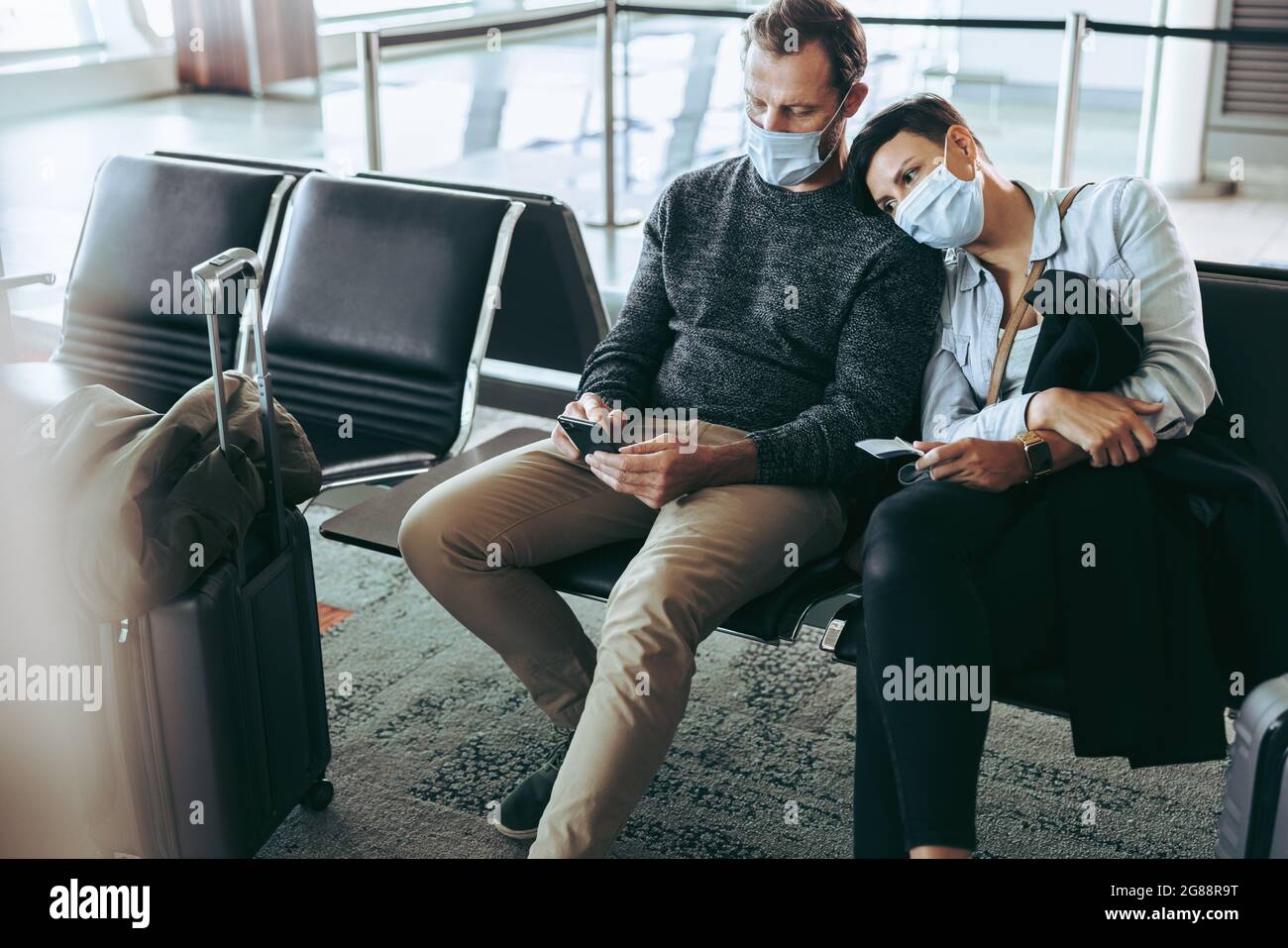 Pärchen mit Gesichtsmasken, die im Wartebereich am Flughafen sitzen. Müde Touristen in Gesichtsmasken auf dem Flughafen warten auf verspäteten Flug wegen Covid-1 Stockfoto