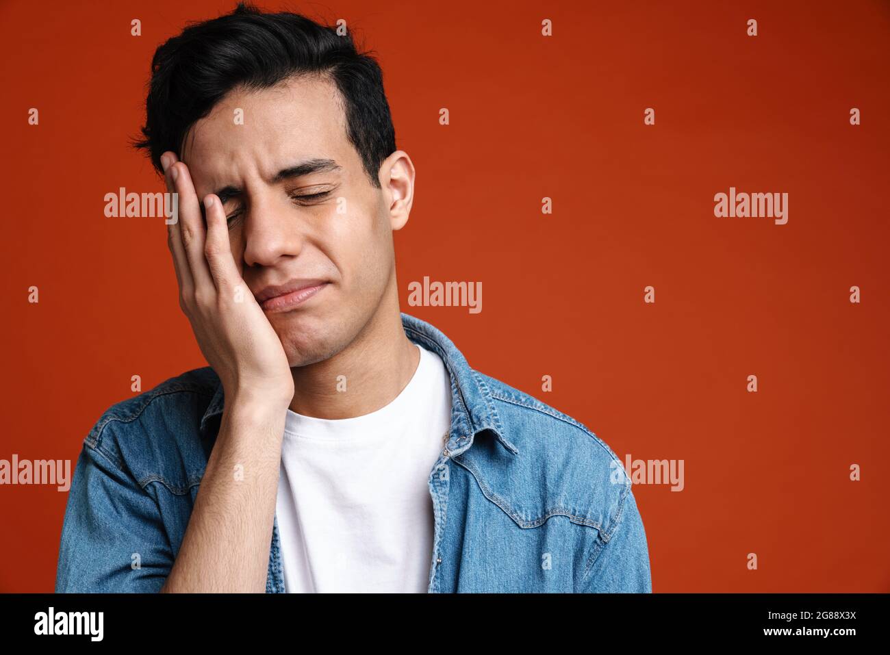 Trauriger hispanischer Mann im Hemd runzelte die Stirn, während er sein Gesicht isoliert auf rotem Hintergrund bedeckte Stockfoto