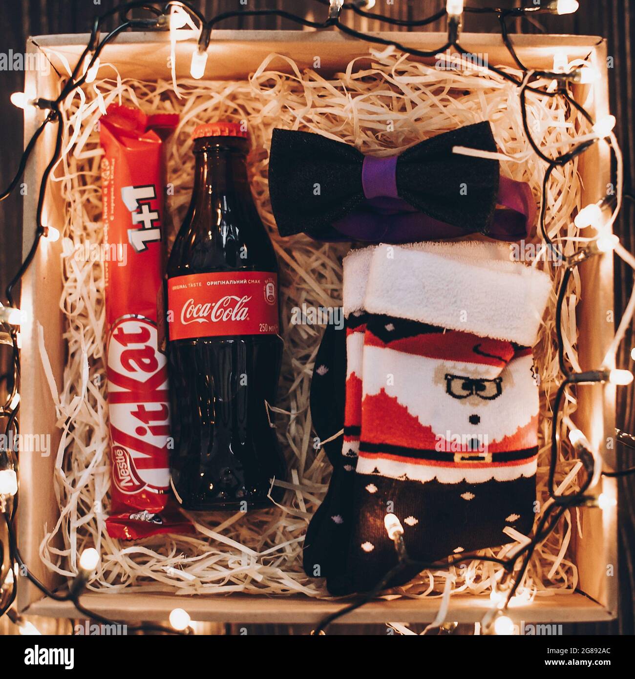 Lviv, Ukraine - 1. Dezember 2017: Coca-Cola-Flasche, stylische Fliege,  weihnachtsmann-Socken und Kit kat on Stroh in Geschenkbox mit  weihnachtsbeleuchtung. Draufsicht. Gif Stockfotografie - Alamy