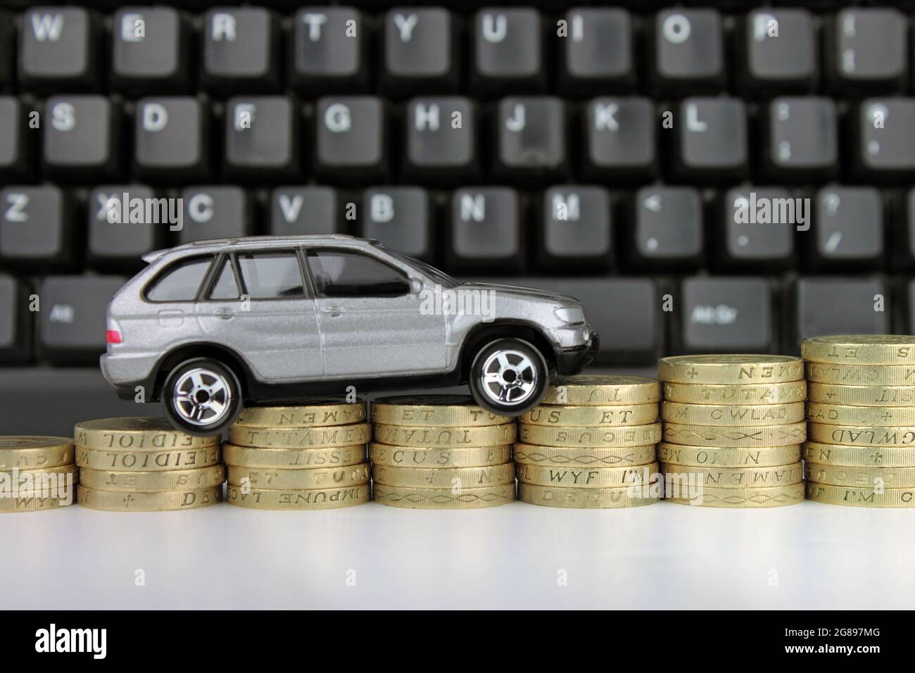 Ein Auto auf einer aufsteigenden Münzsäule mit einer Tastatur im Hintergrund. Eine Metapher für Online-Autokauf, Steuern, Versicherungen und Shopping. Stockfoto