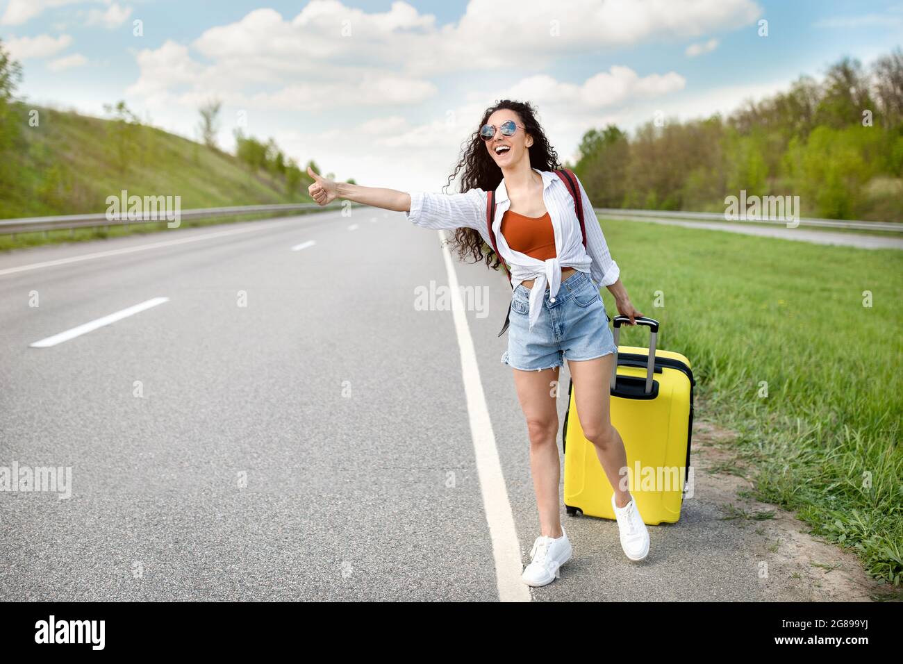 Fröhliche Millennial-Dame, die ihre Fahrt hinkitzeln lässt, Auto mit Koffer auf der Autobahn fängt, auf dem Land unterwegs ist, Platz kopieren Stockfoto