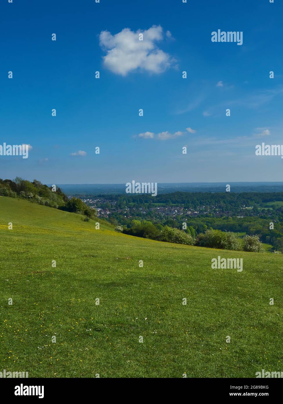 Die geschwungene Stirn eines grasbewachsenen Hügels, mit Blick auf ein grünes Tal mit einer kleinen Stadt. Über dem Kopf hängt ein sommerlicher Himmel mit einer einzelnen markanten Wolke. Stockfoto