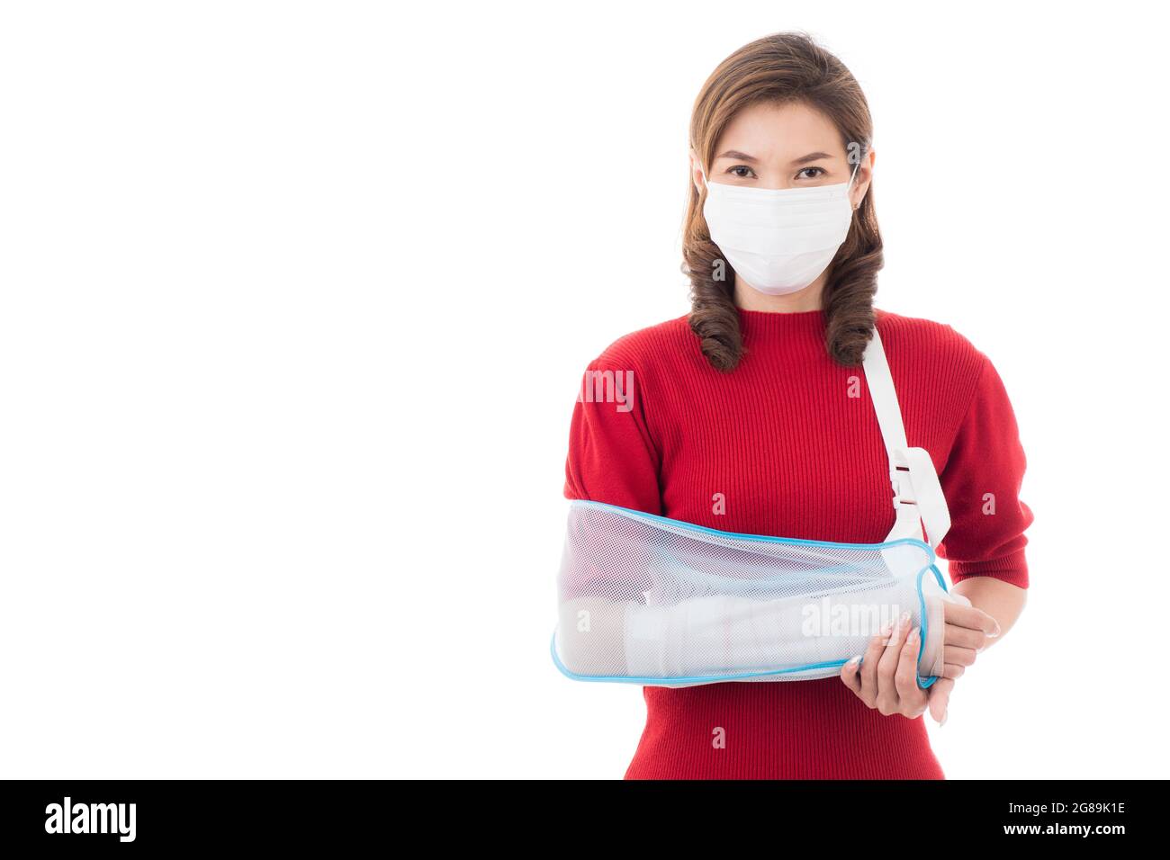 Schöne asiatische Frau mit gebrochenem Arm und Verband tragen chirurgische schützende Hygienemaske stehend isoliert auf weißem Hintergrund mit Smiley Gesicht. Ins Stockfoto