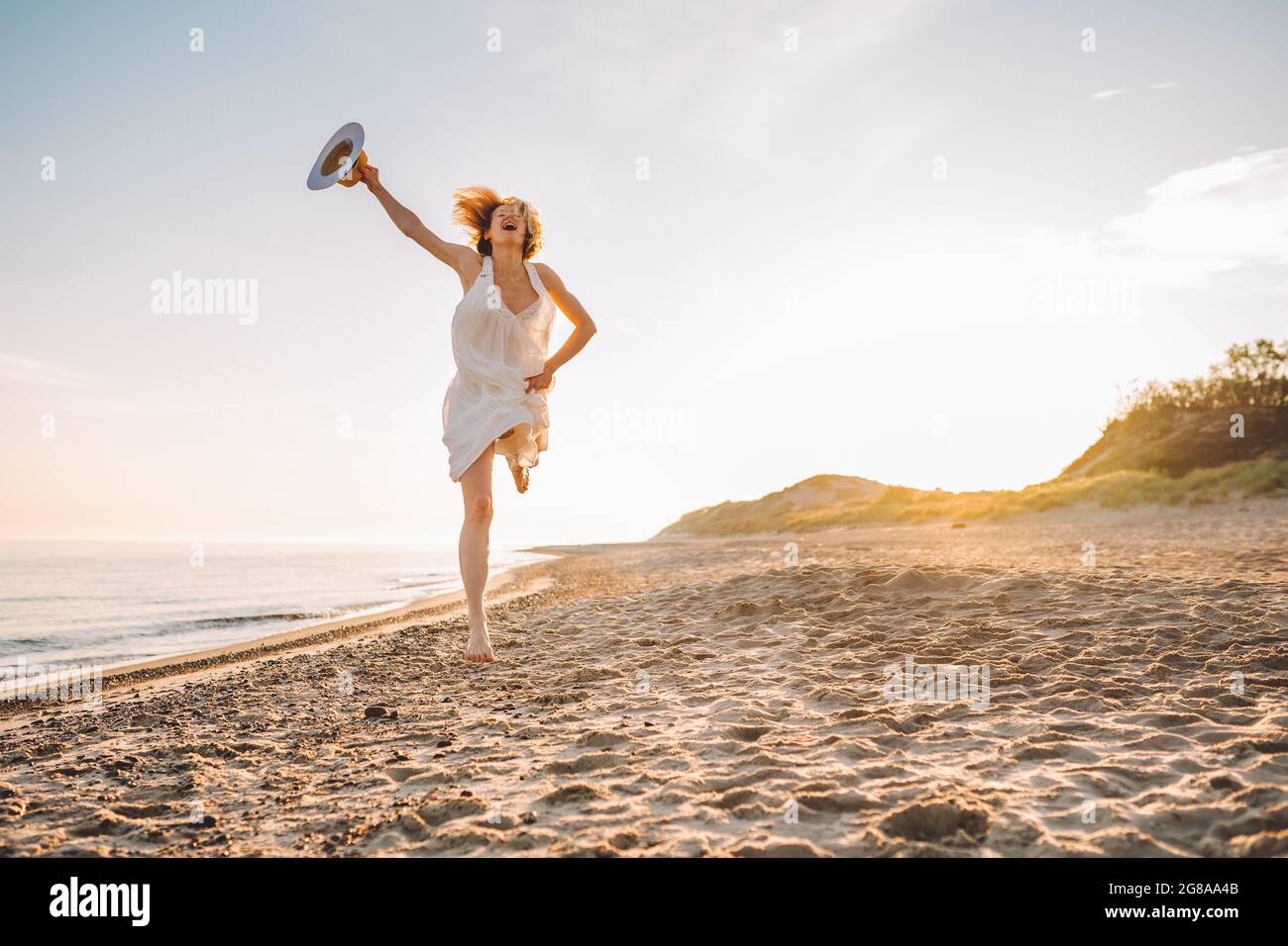 Glückliche junge Frau in weißem, langem Kleid und Hut tanzen und springen in der Dämmerung am Strand. Glück, Freiheit. Stockfoto