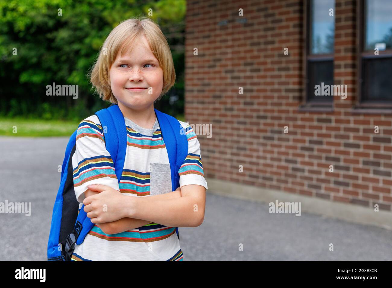 Schönes fröhliches Kind in der Nähe des Schulgebäudes auf dem Schulhof. Kleine blonde Studentin mit blauem Rucksack. Zurück zum Schulkonzept. Stockfoto