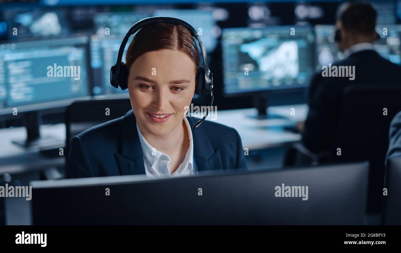 Happy Beautiful Technical Customer Support Specialist spricht an einem Headset, während er an einem Computer in einem dunklen Überwachungs- und Kontrollraum arbeitet Stockfoto