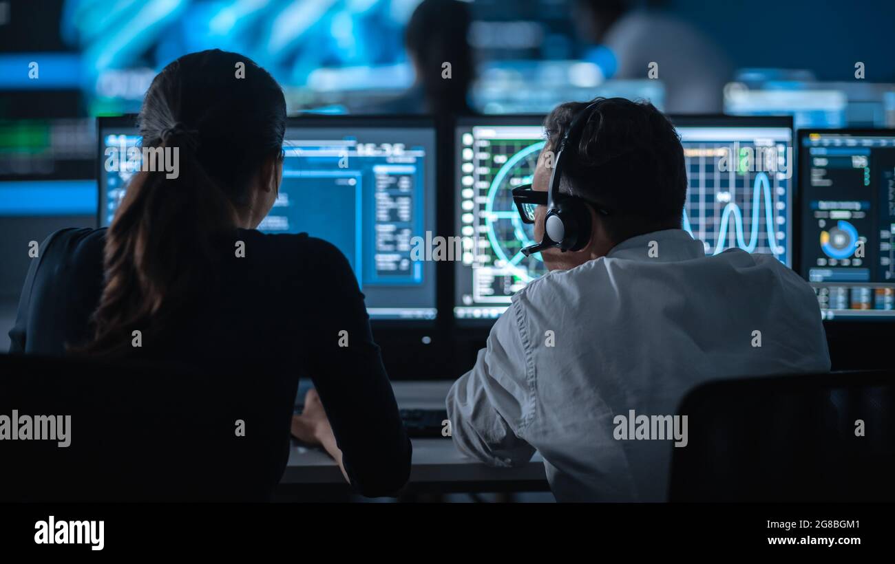 Zwei Spezialisten arbeiten an einem Computer mit Live-Ananlyse-Feed von einer globalen Karte auf einem großen digitalen Bildschirm. Die Mitarbeiter sitzen mit vor den Displays Stockfoto