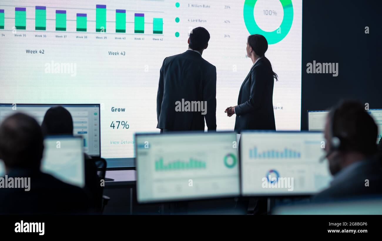 Zwei Händler, die ein Meeting in einem modernen Überwachungsbüro mit Analyse-Feed auf einem großen digitalen Bildschirm haben. Überwachungsraum mit Maklern und Finanzen Stockfoto