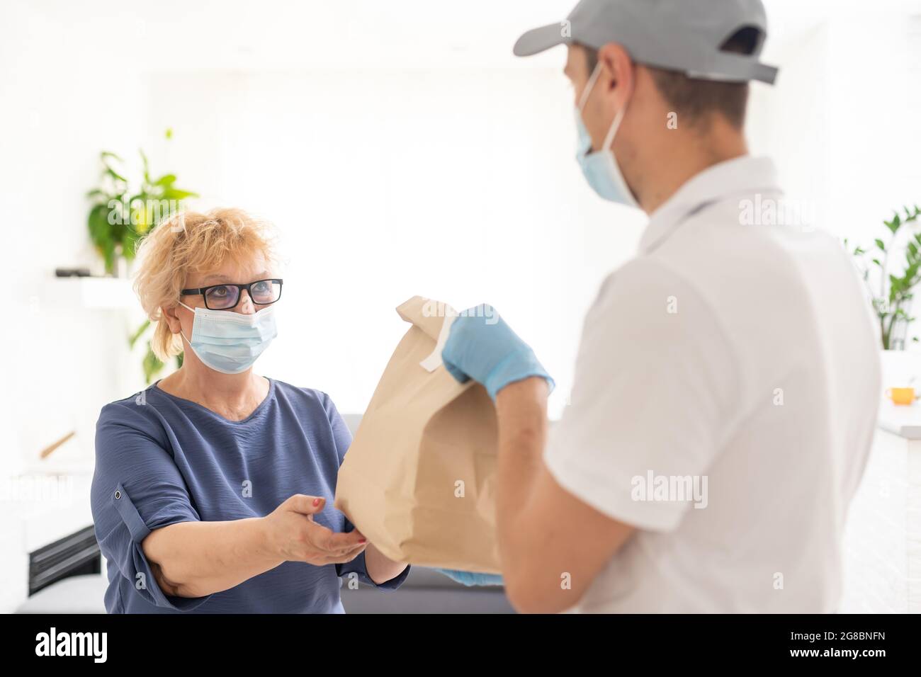 Eine ältere maskierte Frau wird vor der Haustür ihres Hauses ein Paket ausgehändigt Stockfoto