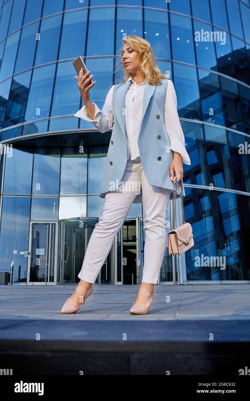 Frau im Business-Anzug steht vor einem Bürogebäude und schaut auf den Smartphone-Bildschirm. Stockfoto