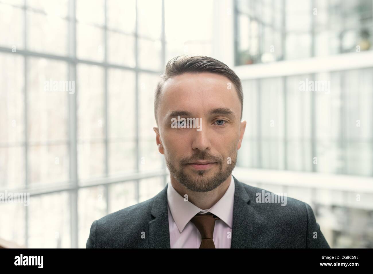 Porträt eines selbstbewussten, eleganten Geschäftsmannes, der die Kamera im Büro mit großen Fenstern im Hintergrund betrachtet Stockfoto