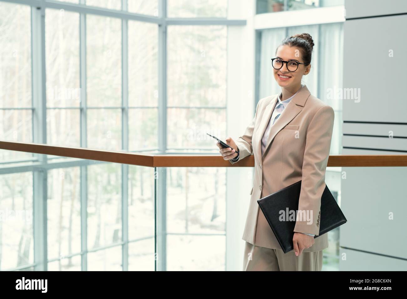 Porträt einer jungen eleganten Geschäftsfrau in Anzug lächelnd auf die Kamera, während sie auf dem Büroflur mit Ordner stand und ihr Mobiltelefon benutzte Stockfoto