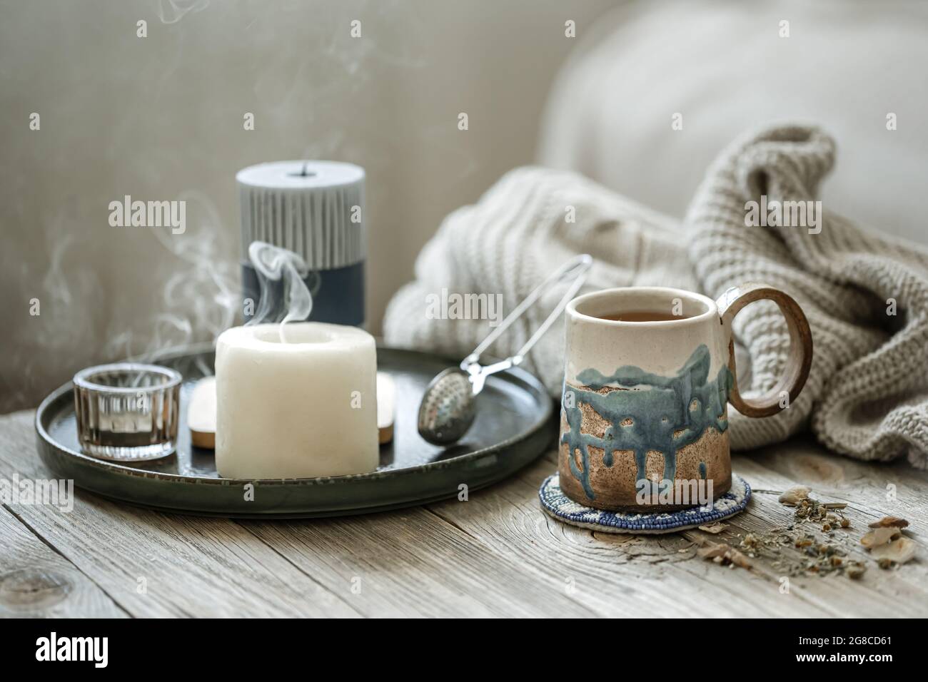 Gemütliche Komposition mit Keramikbecher, Kerzen und einem gestrickten Element auf unscharfem Hintergrund. Stockfoto