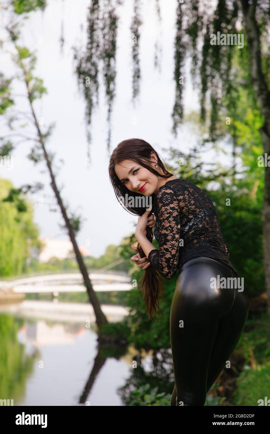 Das hübsche langbeinige Mädchen steht in einem grünen Park in der Nähe des Wassers; sie trägt schwarze Kleidung (transparentes Oberteil und glänzende enge Leggings). Sie dreht sich um und schaut uns an. Stockfoto