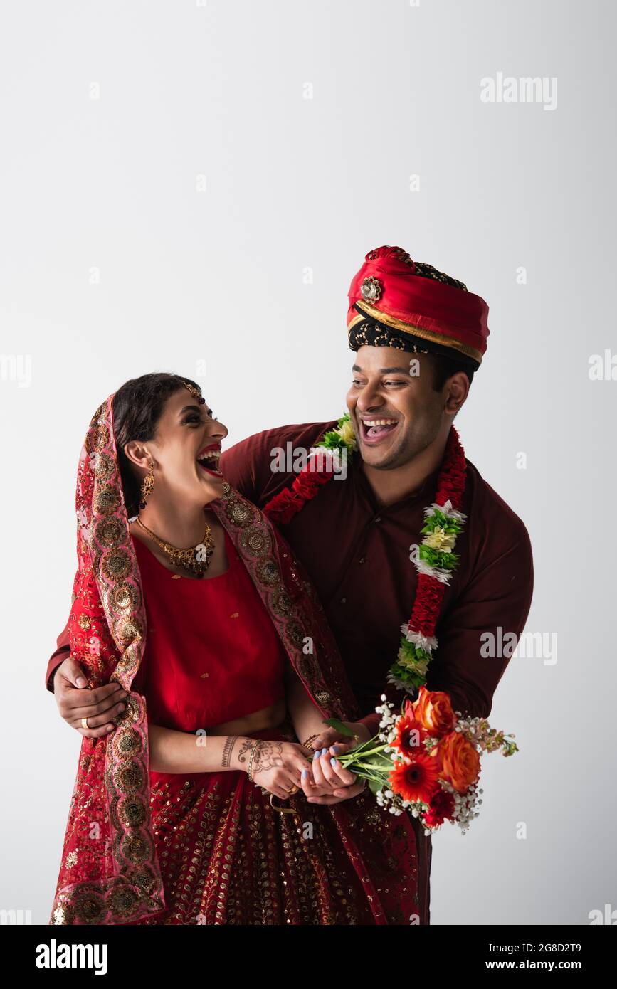 Glücklicher indischer Bräutigam im Turban umarmt fröhliche Braut in Sari mit Blumen isoliert auf grau Stockfoto