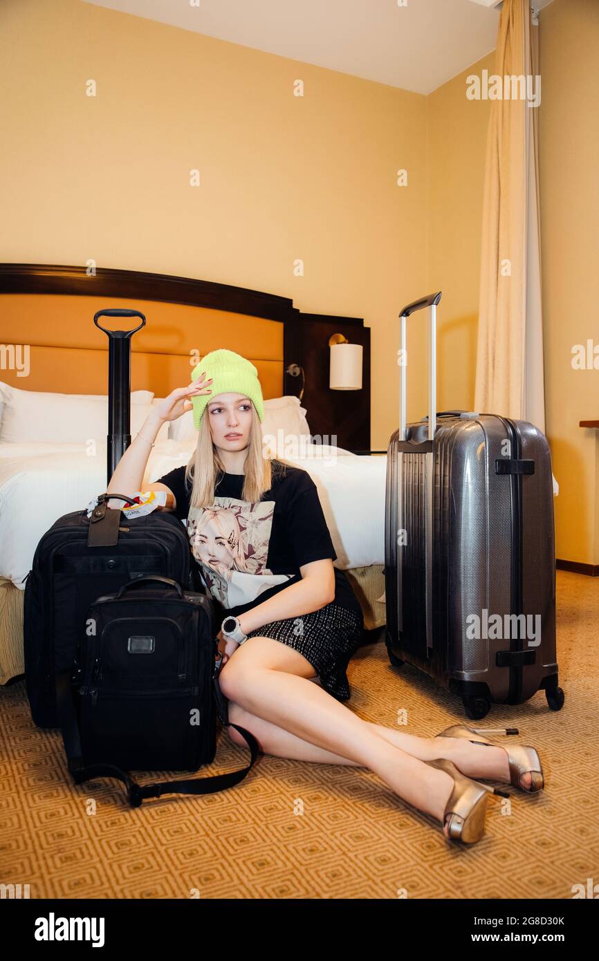 Hübsche blonde Frau, die drinnen Winter- und Sommerkleidung trägt, Hut, T-Shirt, kurzen Rock, sitzt vor einem Bett, möglicherweise in einem Hotel Stockfoto