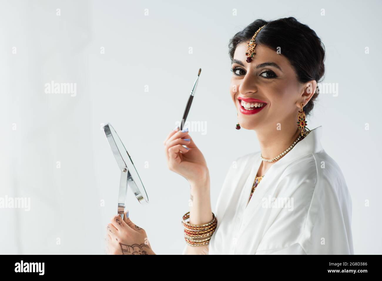 Glückliche indische Braut mit Mehndi hält Spiegel und Kosmetikbürste auf weiß Stockfoto