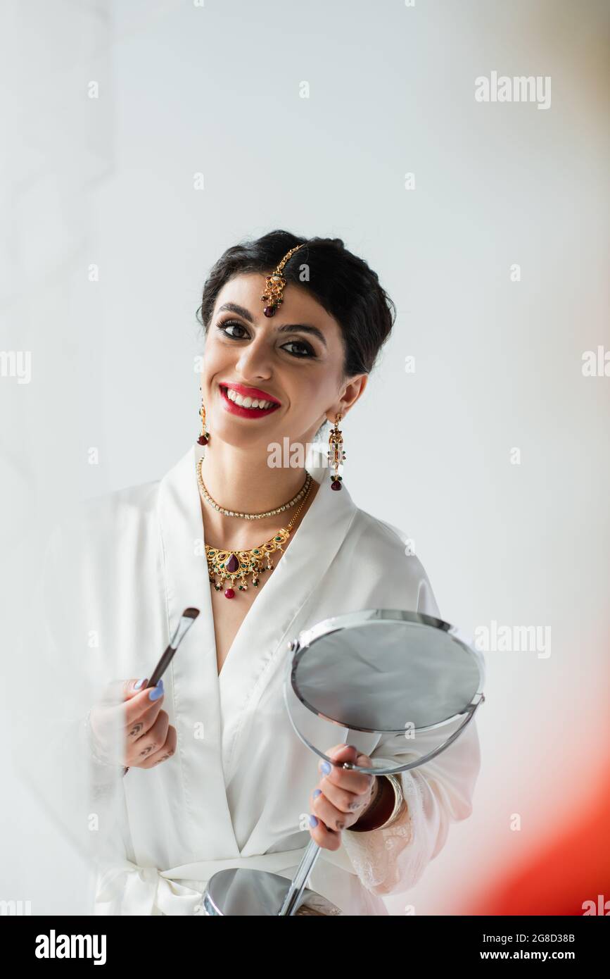 Fröhliche indische Braut mit Mehndi hält Spiegel und Kosmetikbürste auf weiß Stockfoto