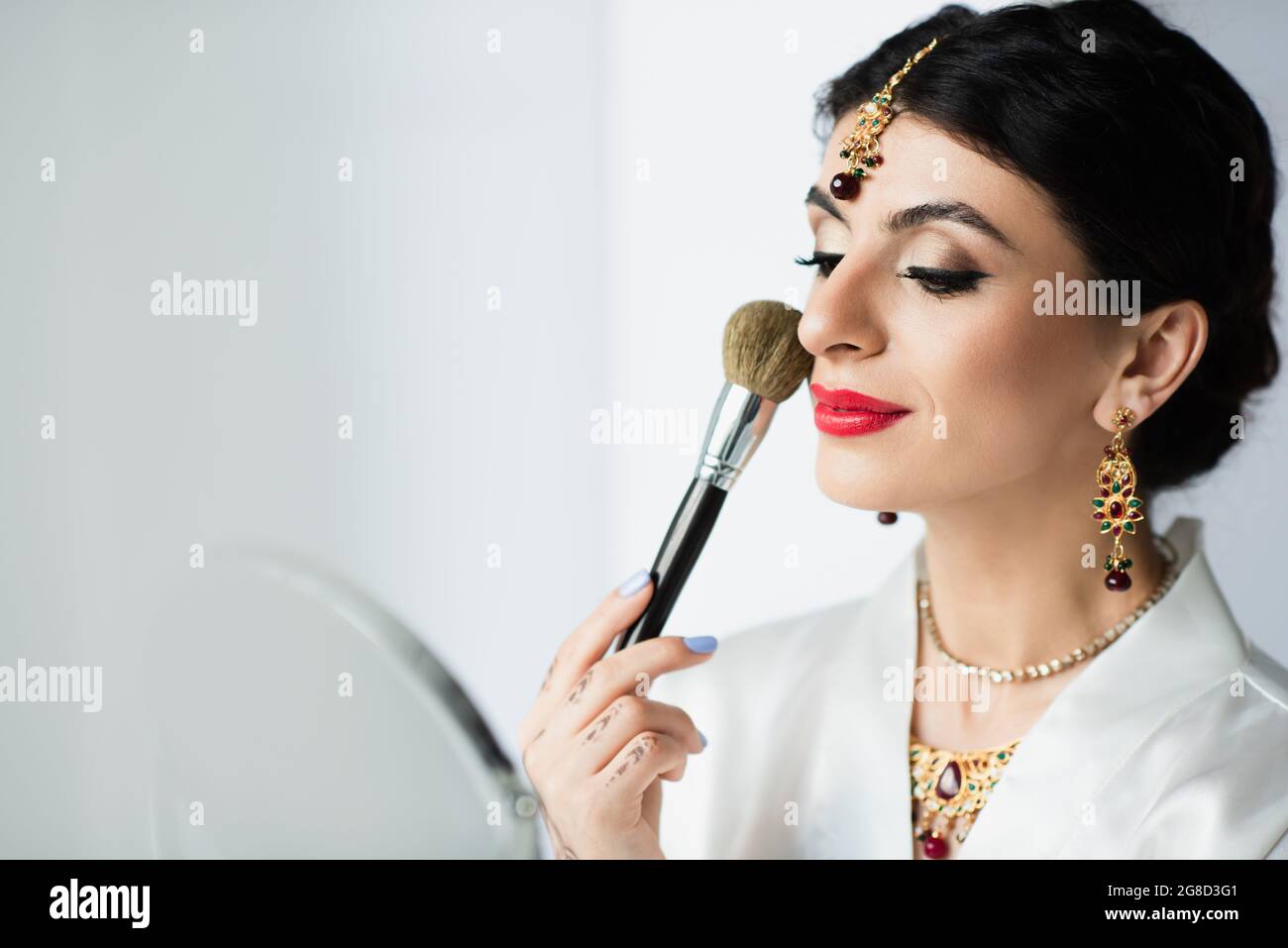 indische Braut mit Mehndi, das Gesichtspuder mit Kosmetikpinsel neben dem Spiegel auf Weiß aufgibt Stockfoto