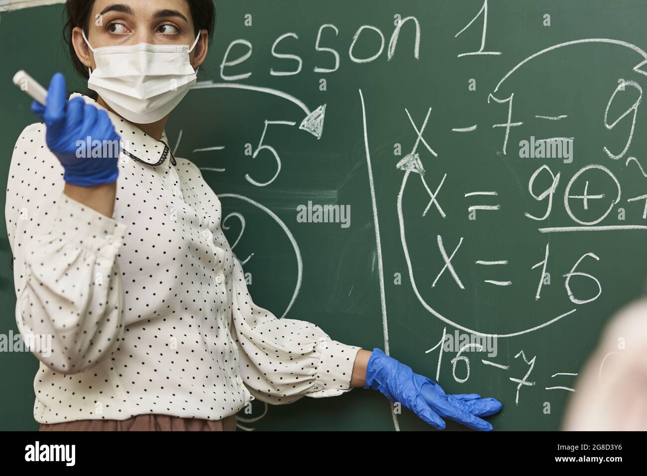 Männlicher Lehrer, der eine Gesichtsmask mit Latexhandschuhen trägt und der Klasse eine mathematische Formel erklärt Stockfoto