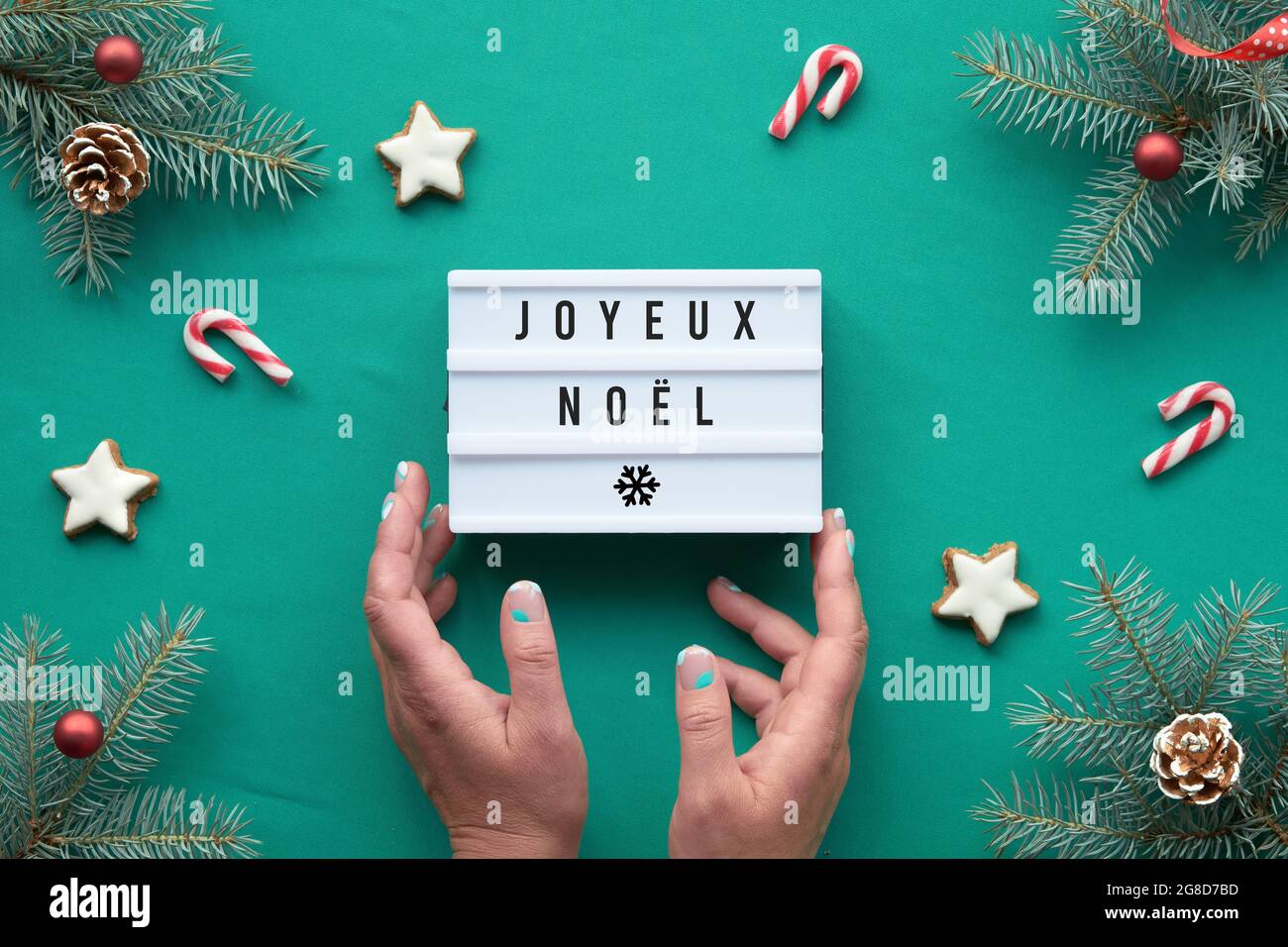 Leuchtkasten mit Text Joyeux Noel - Frohe Weihnachten in französischer Sprache. Hände halten die Box. Weihnachten Hintergrund, Draufsicht auf Tannenzweigen mit roten Verzierungen Stockfoto