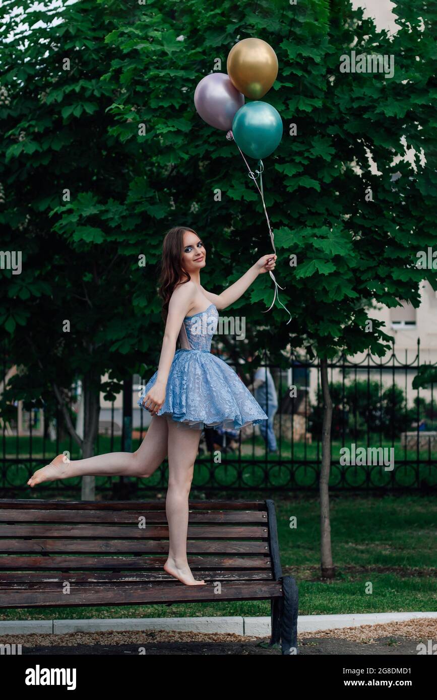 Eine junge Frau in einem blauen Kleid geht barfuß auf einer Holzbank und hält fliegende Ballons, so als ob sie fliegen würde Stockfoto