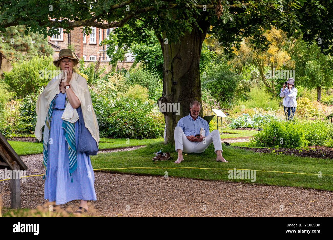 Eine Frau, die verwirrt aussieht, ein Mann, der an einem Baum liest und eine andere Frau, die im Hintergrund im Chelsea Physic Garden in London telefoniert. Stockfoto