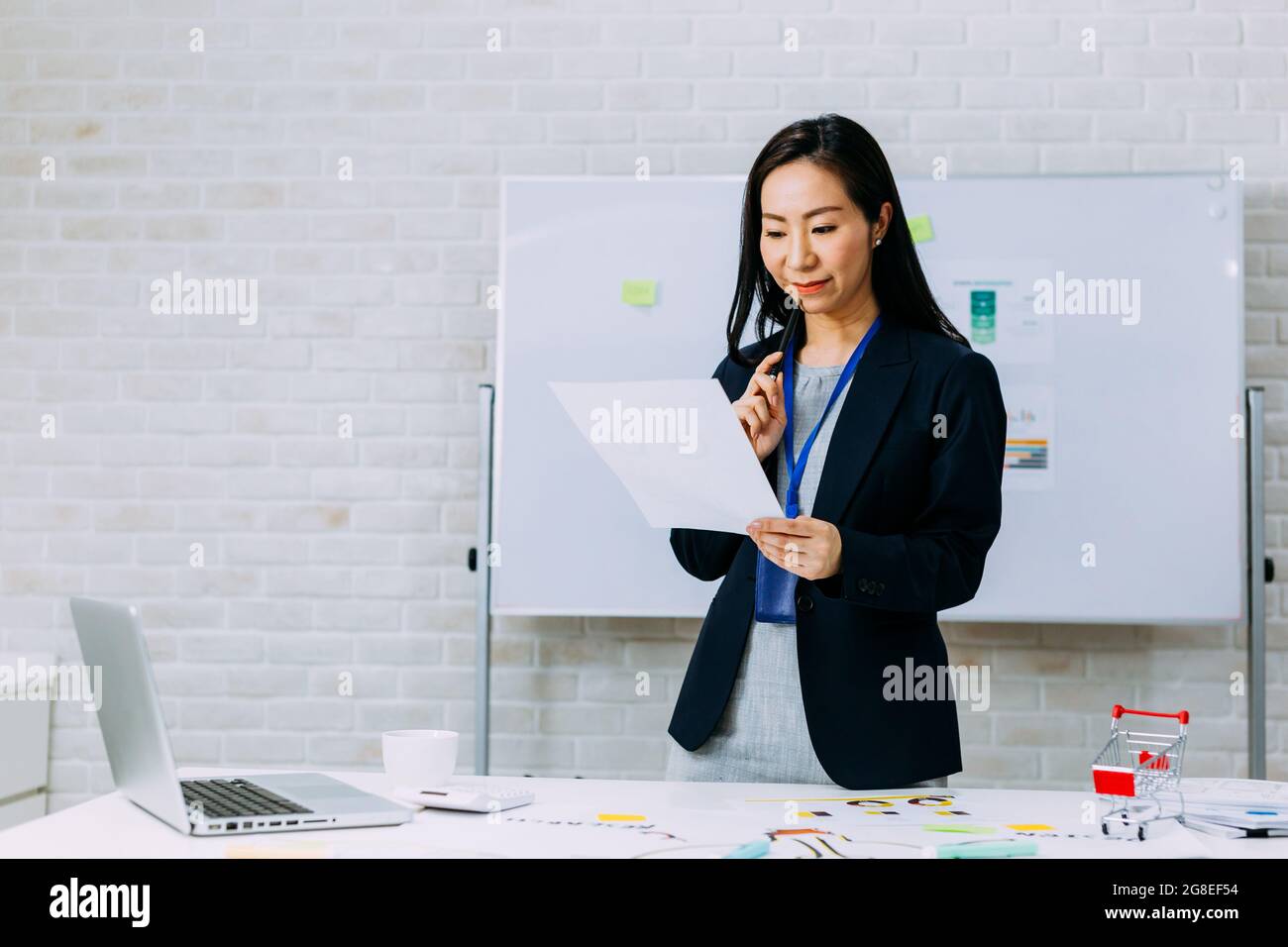 Zufriedene asiatische Frau in formellem Outfit mit Abzeichen lächelnd und Papier zu lesen, während sie am Tisch mit Marketingplan und Laptop im Büro steht Stockfoto