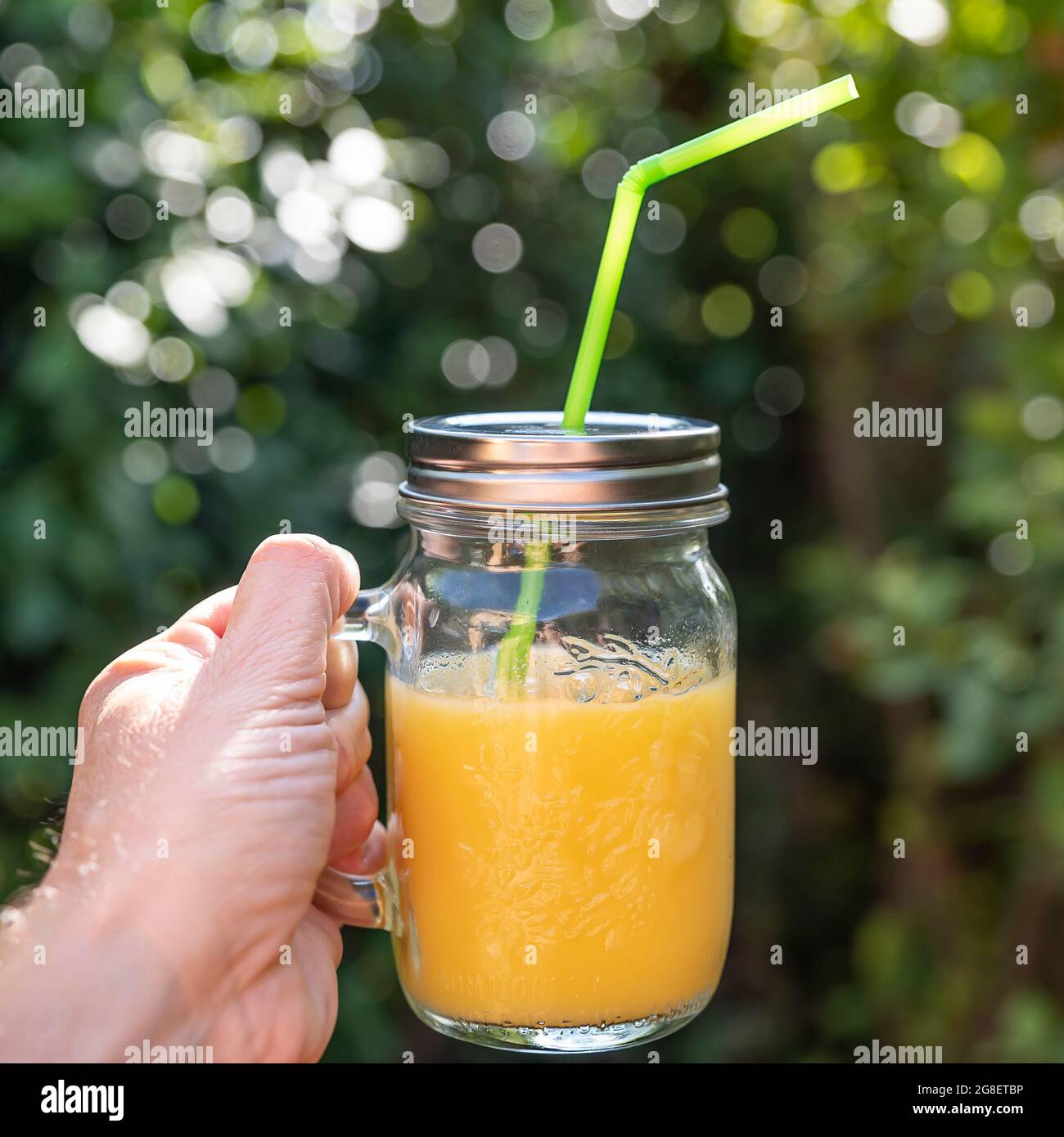 Eine männliche Hand hält ein Glas Fruchtsaft mit einem Stroh auf einem schönen grünen Hintergrund mit Bokeh Stockfoto