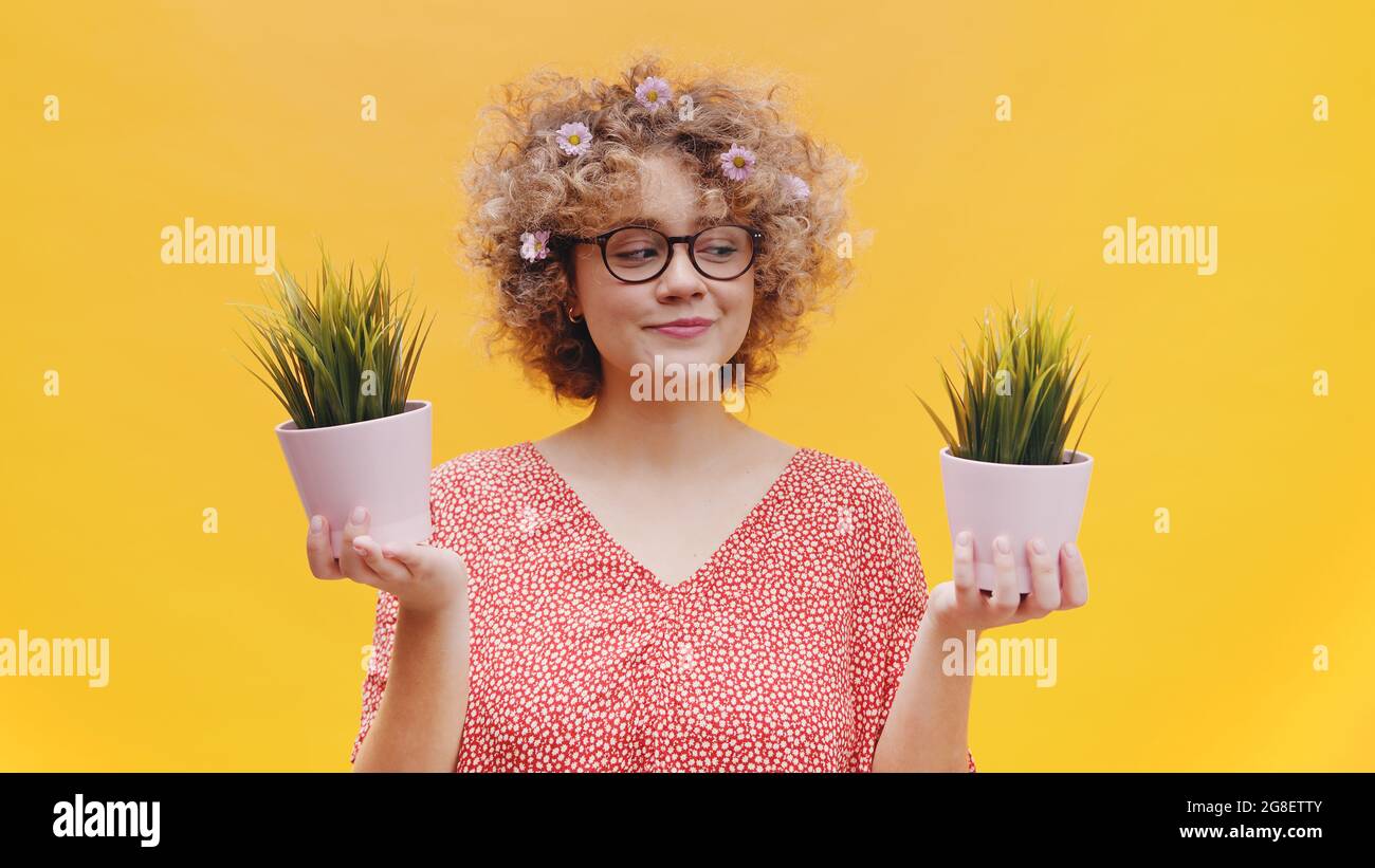 Fröhliches Mädchen, das zwei kleine Topfpflanzen in den Händen hält. Mädchen ist in einem lässigen rosa Top gekleidet und trägt nerdy Brille. Isoliert über hellgelben Hintergrund Studio. Stockfoto