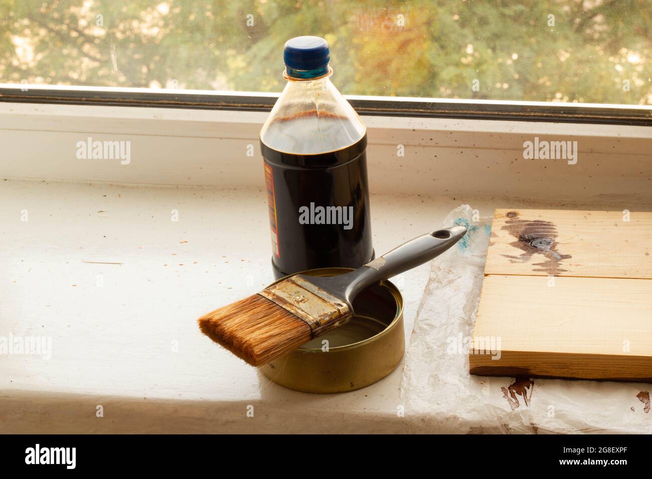 Set zum Bemalen von frisch gehobeltem Holz auf dem Fenster. Eine Flasche brauner Holzfleck und ein Pinsel auf dem Farbbehälter. Grüne Bäume an sonnigen Tagen Stockfoto
