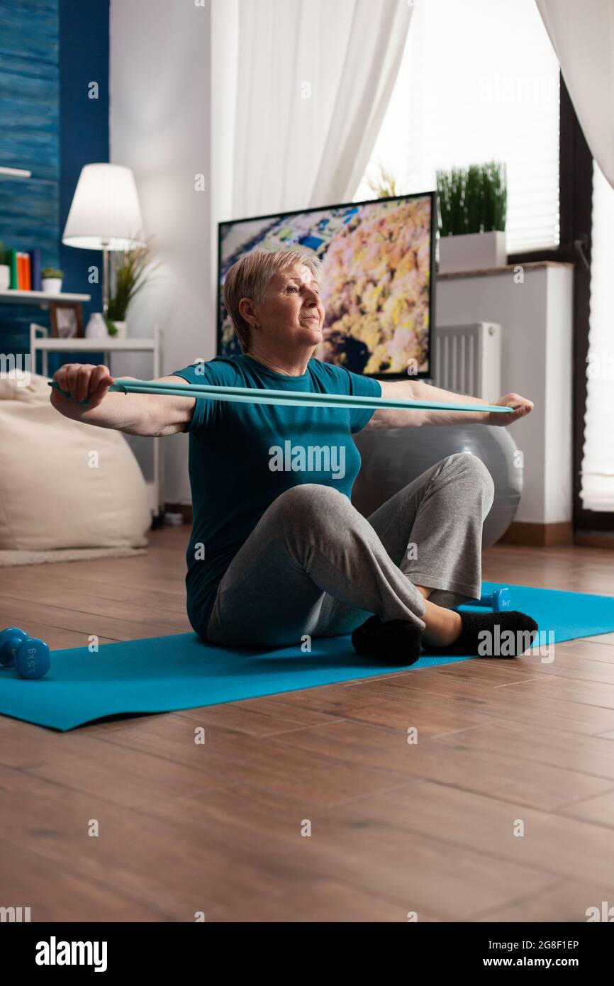 Seniorin im Ruhestand sitzt auf Yoga-Matte Stretching Beine Muskeln mit elastischem Band Training Körperwiderstand. Rentner in Sportbekleidung abnehmen Gewicht beim Muskeltraining im Wohnzimmer Stockfoto