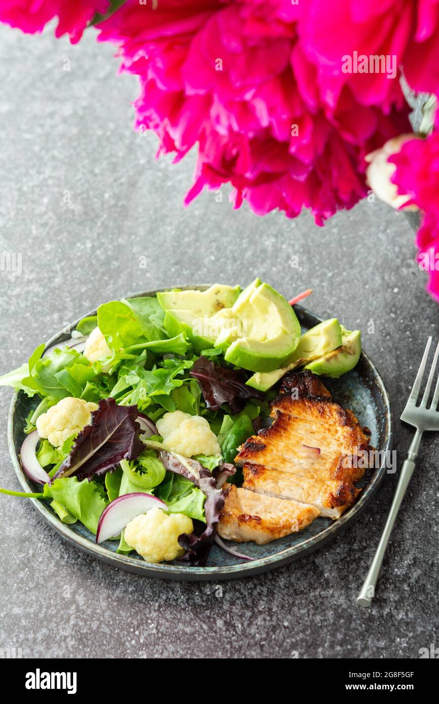 Pfingstrosen in einer Vase und Abendessen auf dem Tisch, ein Teller gebratenes Schweinesteak und ein gemischter Salat mit Blumenkohl und Avocado Stockfoto