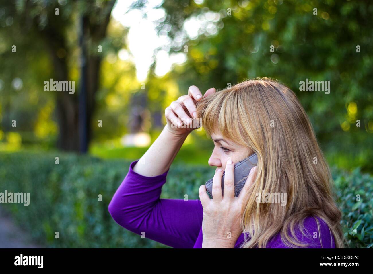 Unschärfe kaukasisch blonde Frau sprechen auf dem Telefon draußen, draußen. 40-jährige Frau in lila Bluse im Park berühren ihre Haare. Ernsthafte Person, die verwendet Stockfoto