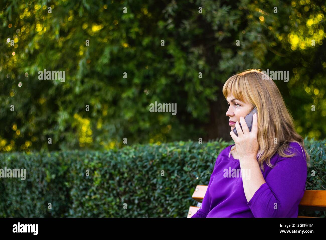 Unschärfe kaukasisch blonde Frau reden, sprechen am Telefon draußen, im Freien. 40-jährige Frau in lila Bluse im Park auf der Bank. Erwachsene Person usi Stockfoto