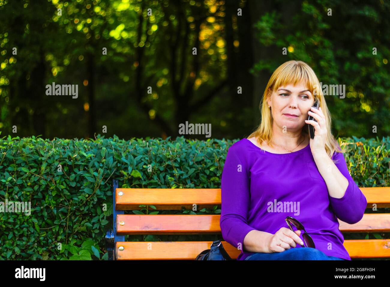 Unschärfe kaukasisch blonde Frau reden, sprechen am Telefon draußen, im Freien. 40-jährige Frau in lila Bluse im Park auf der Bank. Erwachsene, weiblich pro Stockfoto