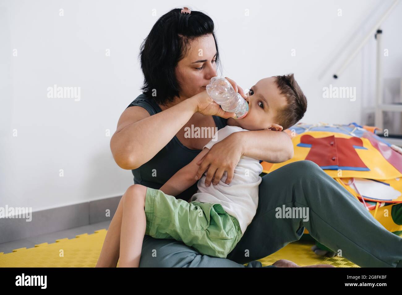 Mutter ernährt sich und gibt behinderten Kindern mit zerebraler Lähmung während der Übungen Trinkwasser. Rehabilitation in der Familie. Stockfoto