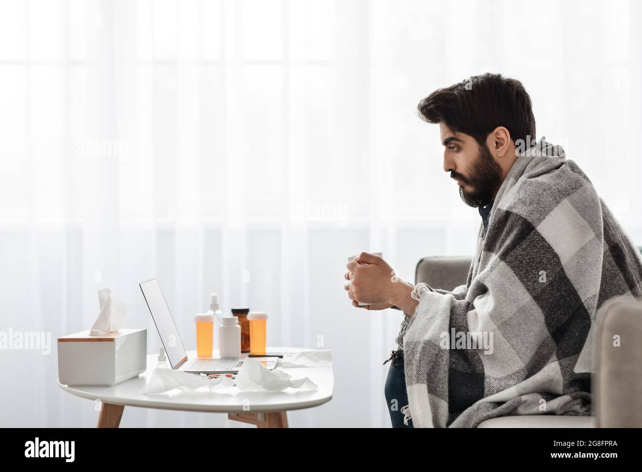 Zu Hause krank. Junger arabischer Mann, der Fieber hat und heißen Tee trinkt, auf dem Sofa sitzt und sich einen Film auf dem Laptop ansieht, Seitenansicht Stockfoto