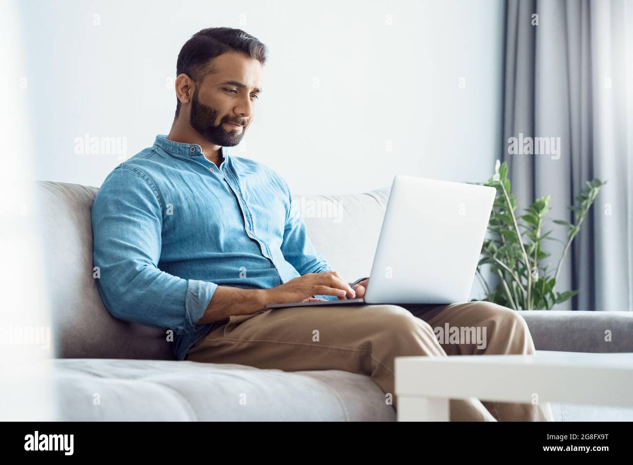 Ernsthafter junger Mann aus dem Osten, der am Laptop arbeitet, während er zu Hause im Hotel auf dem Sofa sitzt Stockfoto