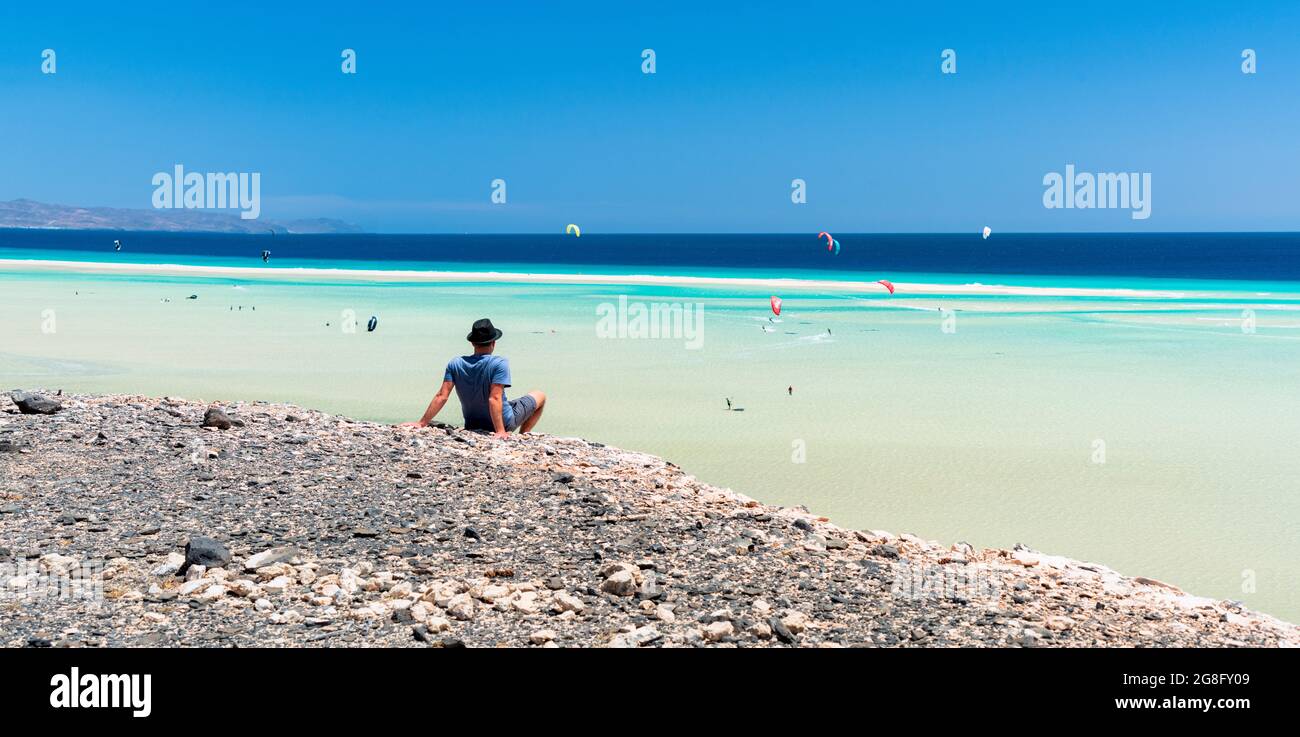 Mann, der auf Felsen sitzt und die Kitesurfer im klaren Meer, Sotavento Beach, Jandia, Fuerteventura, Kanarische Inseln, Spanien, Atlantik, Europa Stockfoto