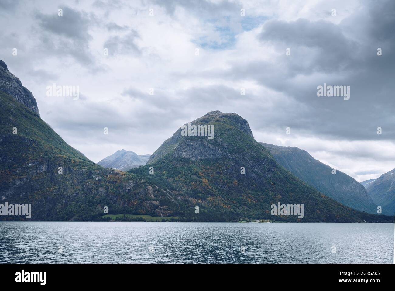 Majestätische Berge ragen an einem bewölkten Tag im arktischen Sommer über den See in Nordnorwegen. Kleine Häuser zwischen den Bergen weit in der Ferne Stockfoto
