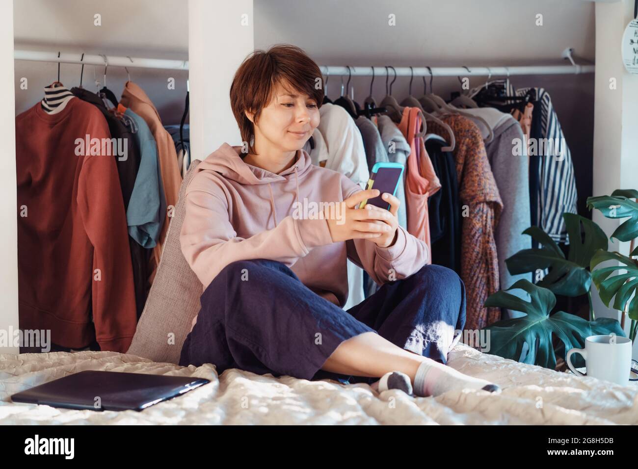 Frau sitzt auf dem Bett und benutzt ihr Smartphone in einer Wohnung mit offenem Kleiderschrank. Gemütliches Zuhause. Online-Kommunikation. Remote-Arbeit. Bildlauf Stockfoto