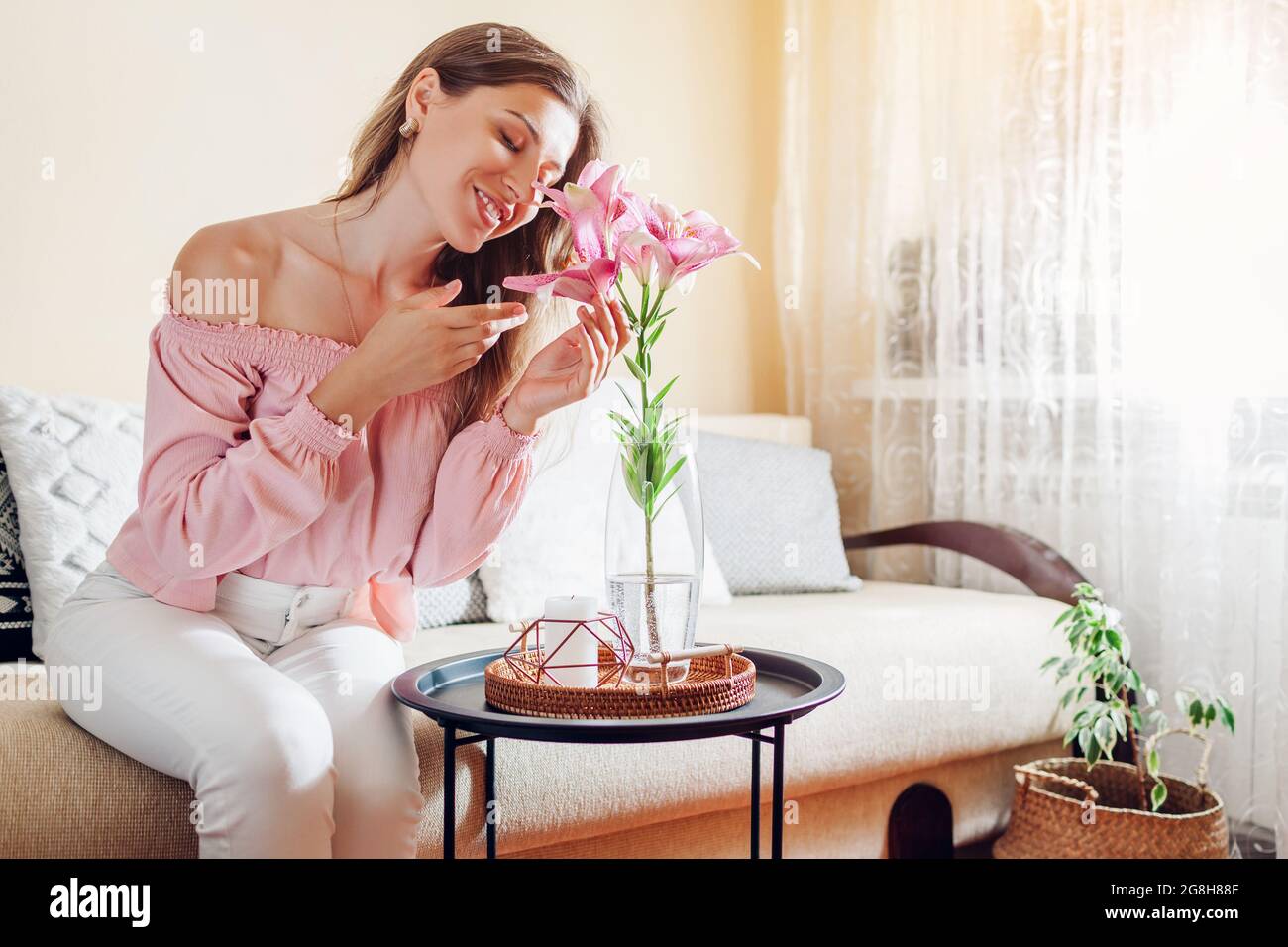Frau riecht rosa Lilie Blumen in Vase zu Hause. Junges Modell genießt Bouquet von frischen Blüten. Einrichtung und Einrichtung des Wohnzimmers Stockfoto