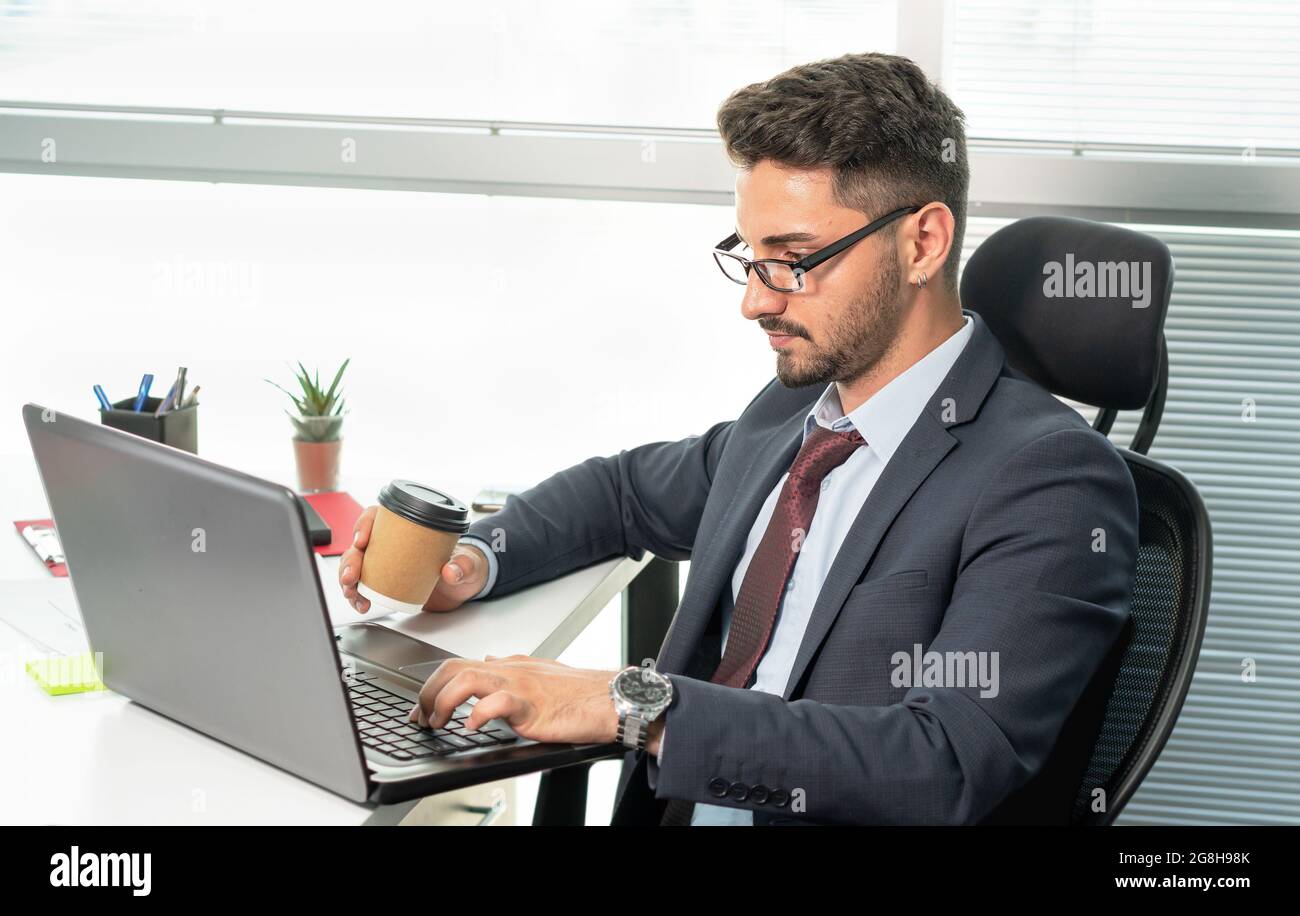 Porträt eines glücklichen Geschäftsmannes in formeller Kleidung, der im Büro mit einem Laptop arbeitet. Fröhlicher, entspannter Mann im Anzug, der beim Kaffee- oder Teetrinken arbeitet. Stockfoto