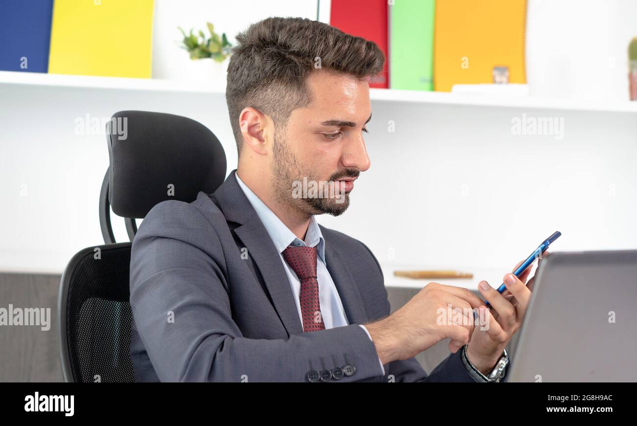 Junger professioneller Geschäftsmann in formeller Kleidung mit Smartphone, während er im modernen Büro am Schreibtisch sitzt. Stockfoto