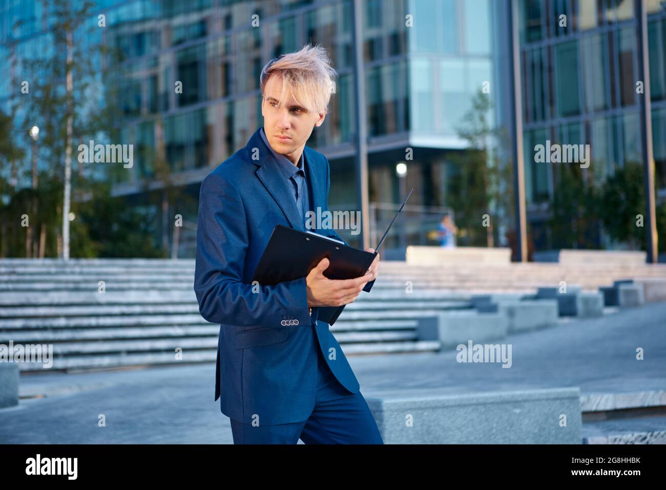 Vor einem Bürogebäude steht ein junger Mann im Geschäftsanzug mit einem Papierordner in der Hand. Stockfoto