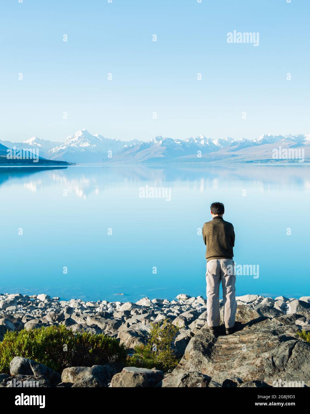 Mann, der am Ufer des Pukaki-Sees steht, tief in Gedanken, mit Mt Cook, der sich im klaren Wasser in der Ferne, South Island, widerspiegelt. Vertikales Format Stockfoto