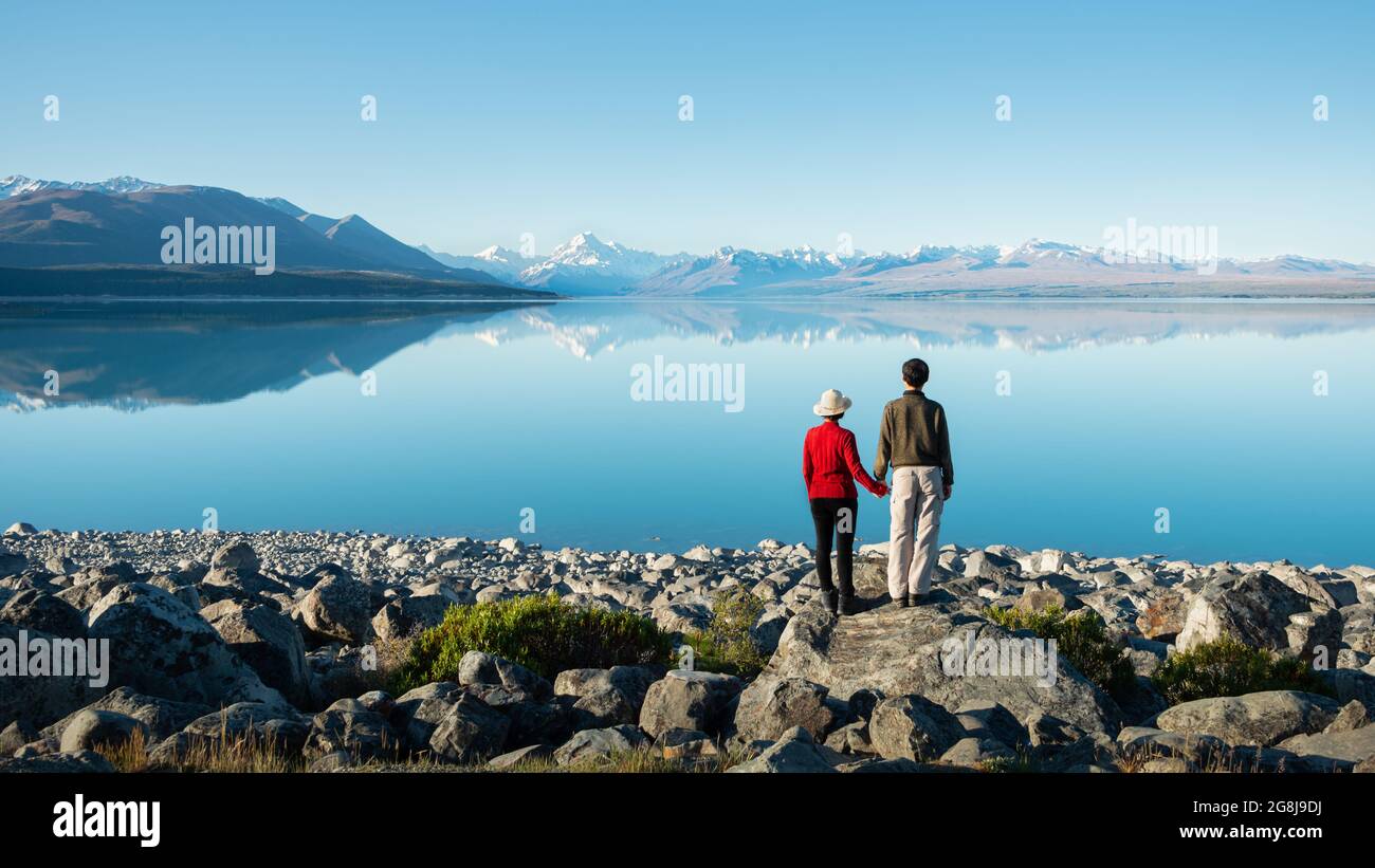 Paar, das am Ufer des Lake Pukaki steht, die Hände hält und den Mt Cook im klaren Wasser, South Island, Neuseeland, beobachten kann Stockfoto
