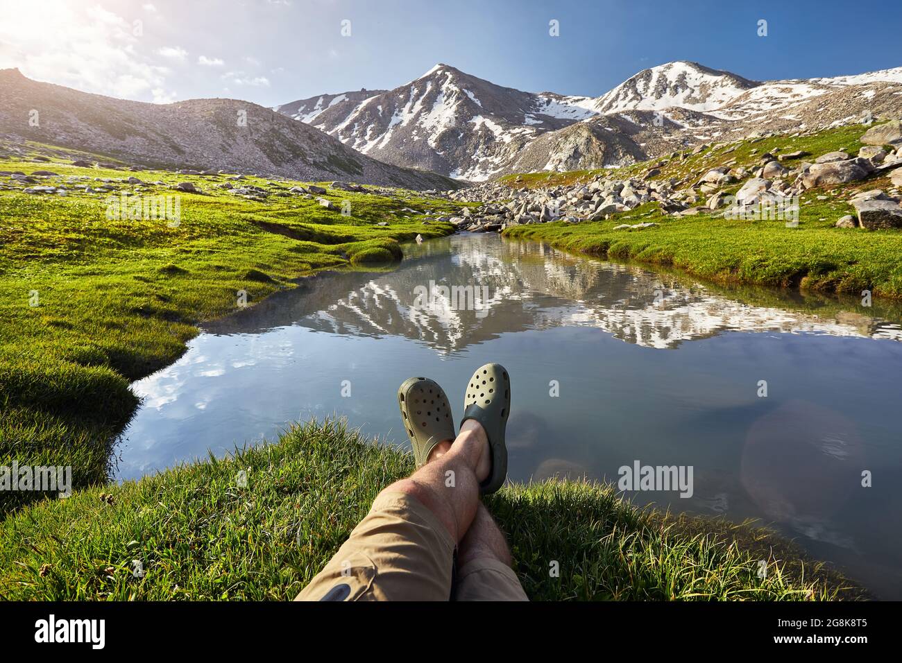 Die Beine des Wanderers in grünen Schuhen ruhen in der Nähe des klaren Bergsees mit Spiegelung des Gipfels mit Schneekappe Stockfoto