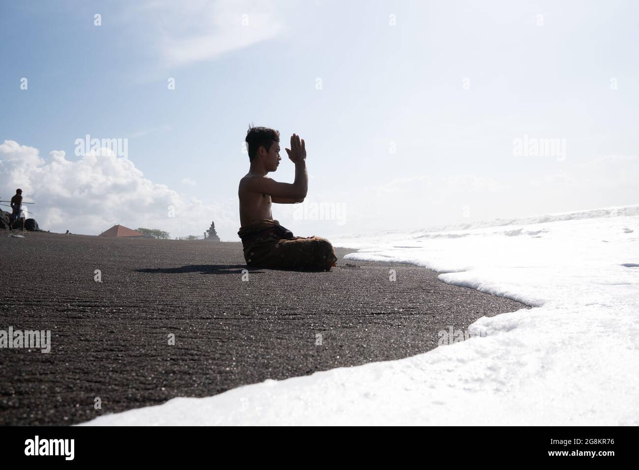 Ein balinesischer Hindu-Mann, der an einem heißen, sonnigen Tag am Strand betet. Der Sand ist schwarz und die Wellen sind schaumig weiß Stockfoto